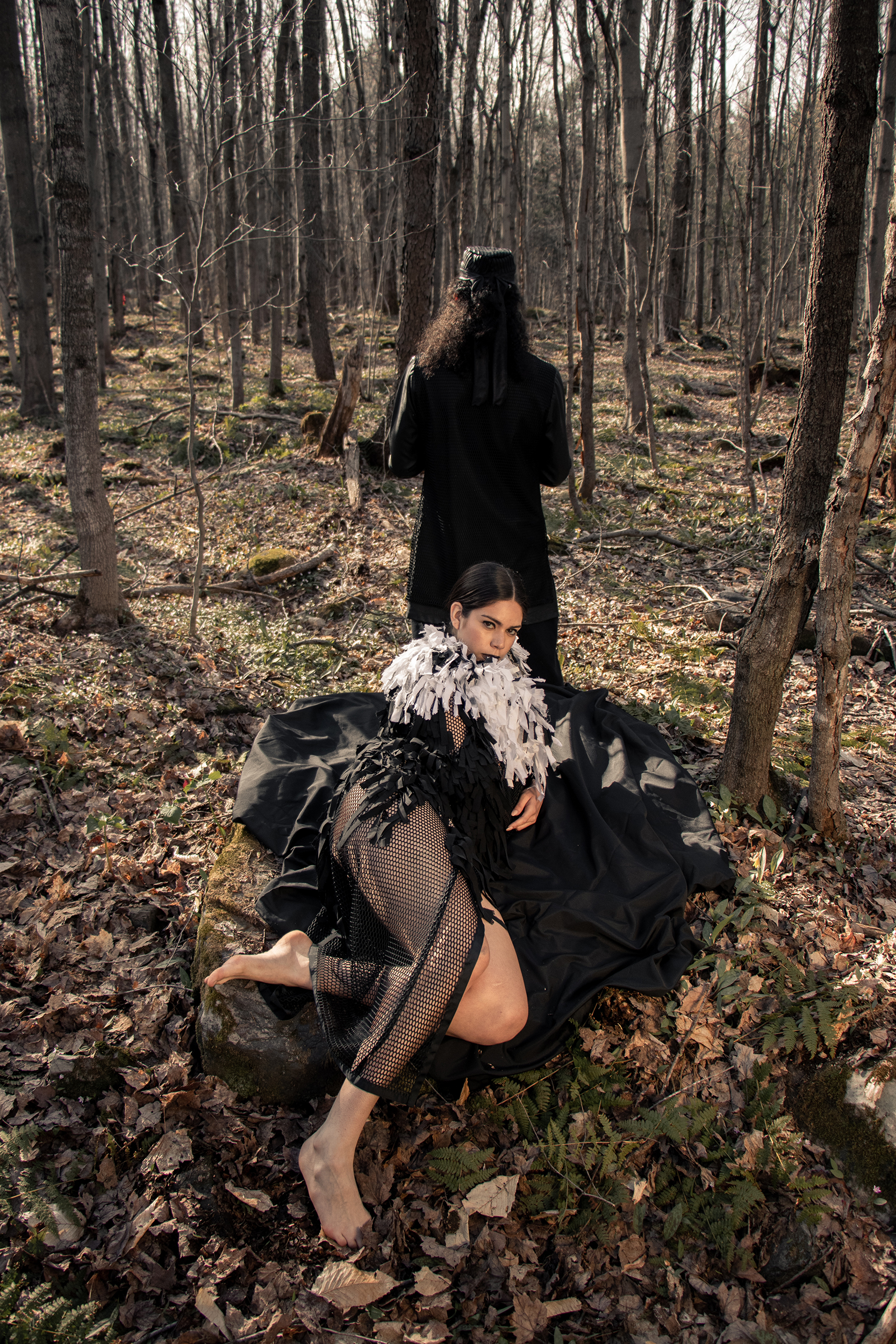 Woman laying on rock in the woods wearing a zero waste black and white fringed cape with man standing in the background