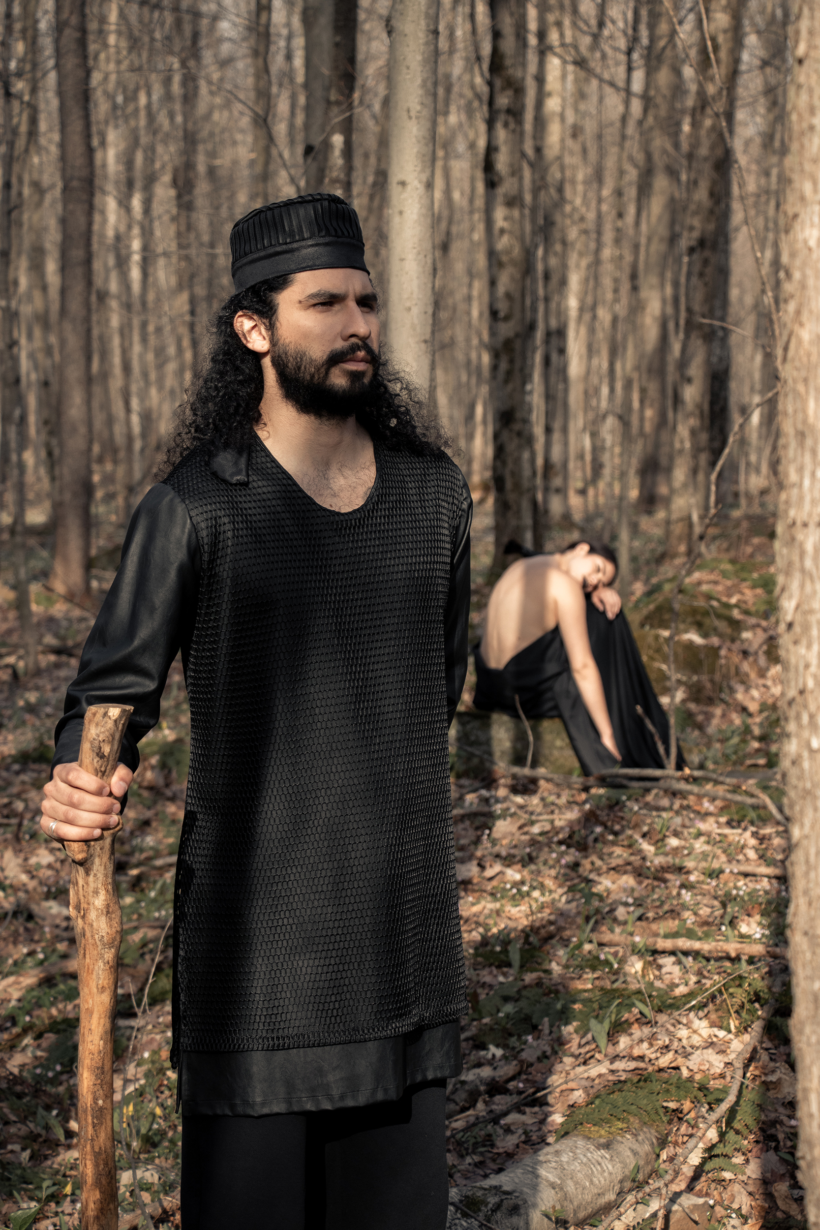 Man wearing black tunic and organic black sweatpants in the woods with woman in the background